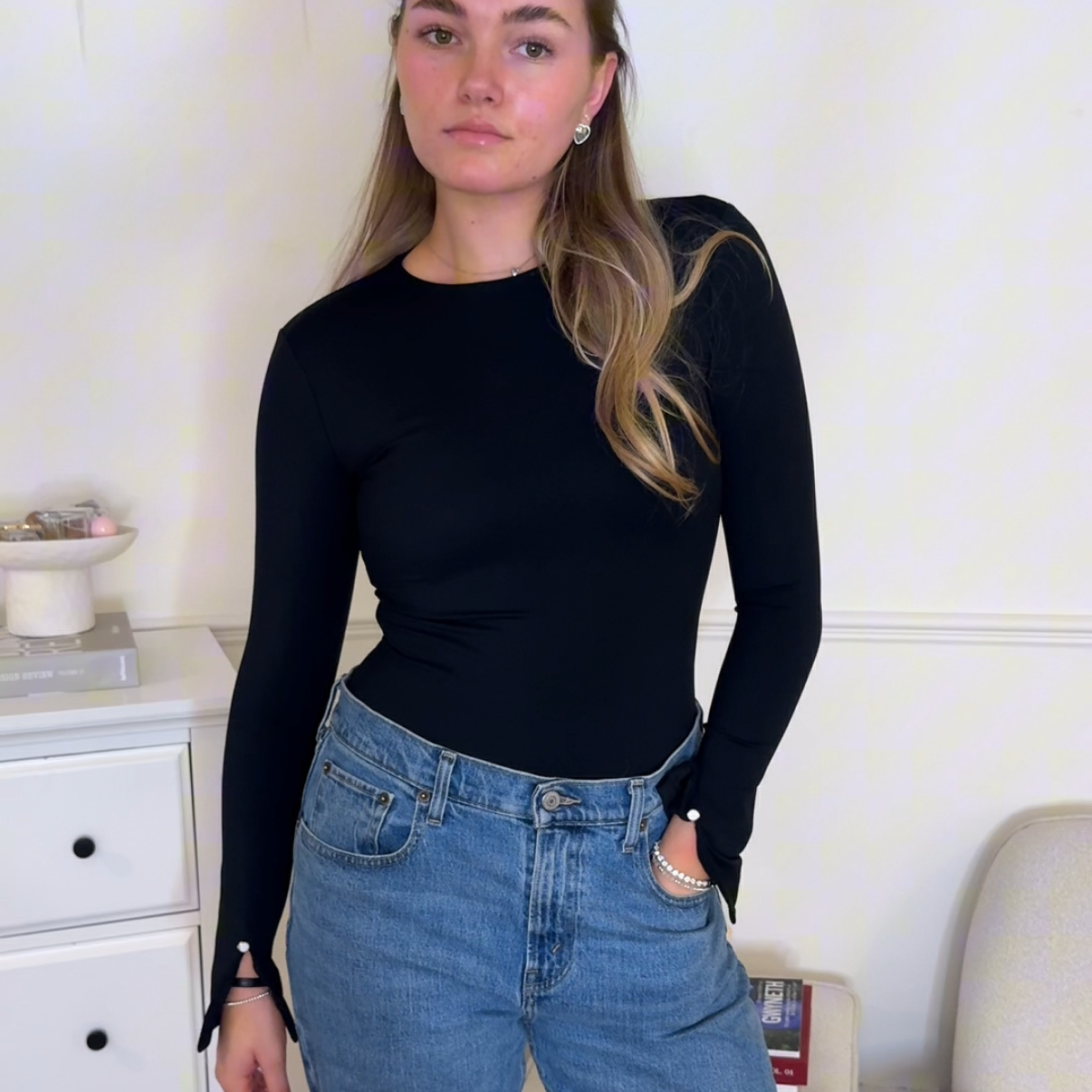Woman wearing a black Ashly Rae top and blue jeans standing in a room with white walls and furniture.