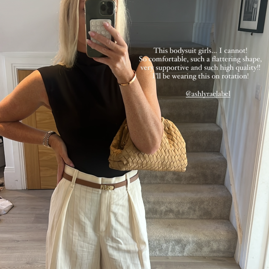 Woman taking a mirror selfie wearing a elegant black top, white shorts, and beige loafers in a home setting.