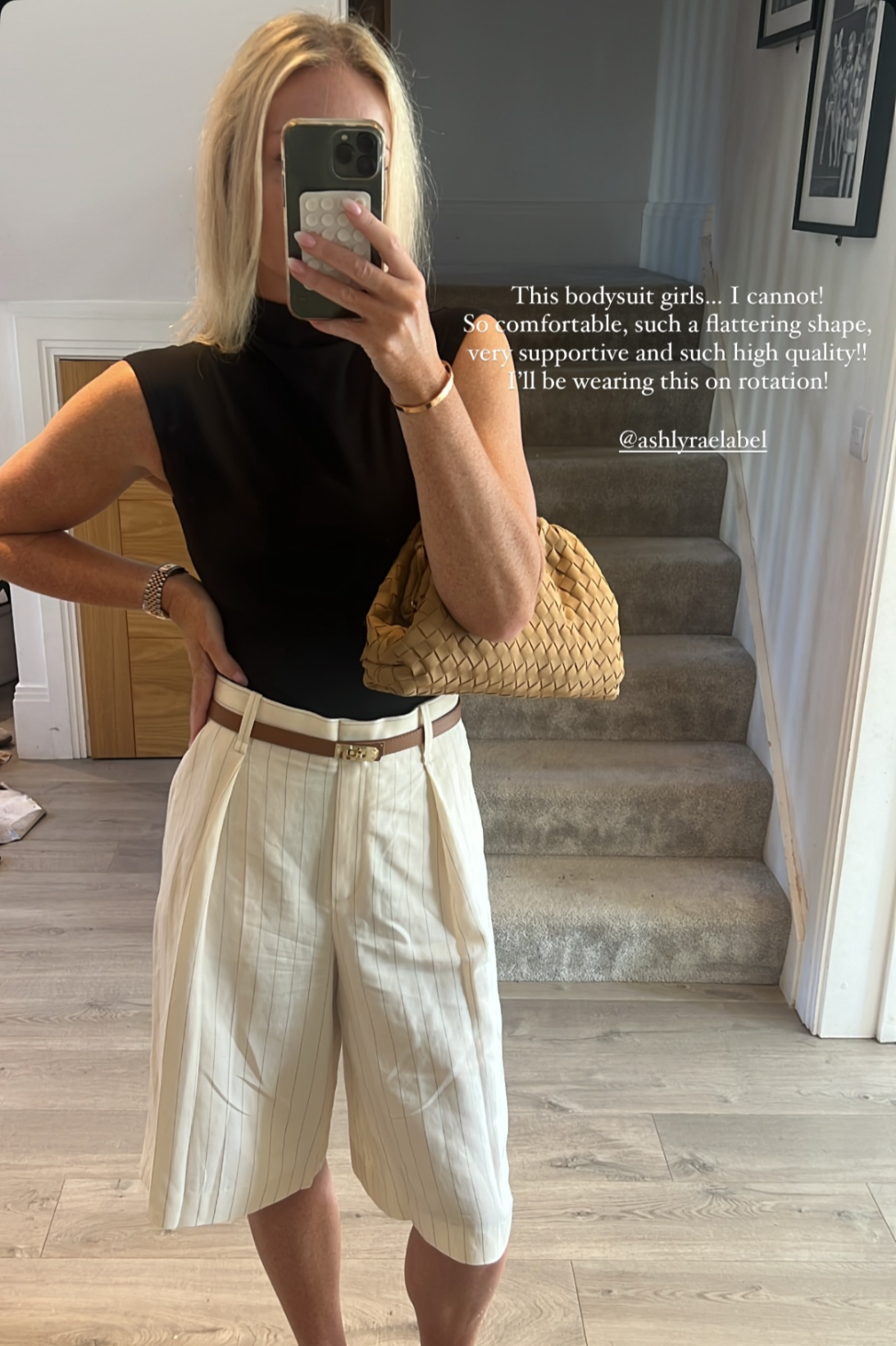 Woman taking a mirror selfie wearing a elegant black top, white shorts, and beige loafers in a home setting.
