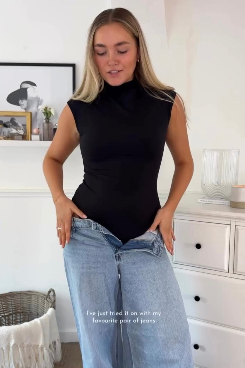 Woman wearing a black sleeveless top and light blue jeans in a room with white walls and furniture.