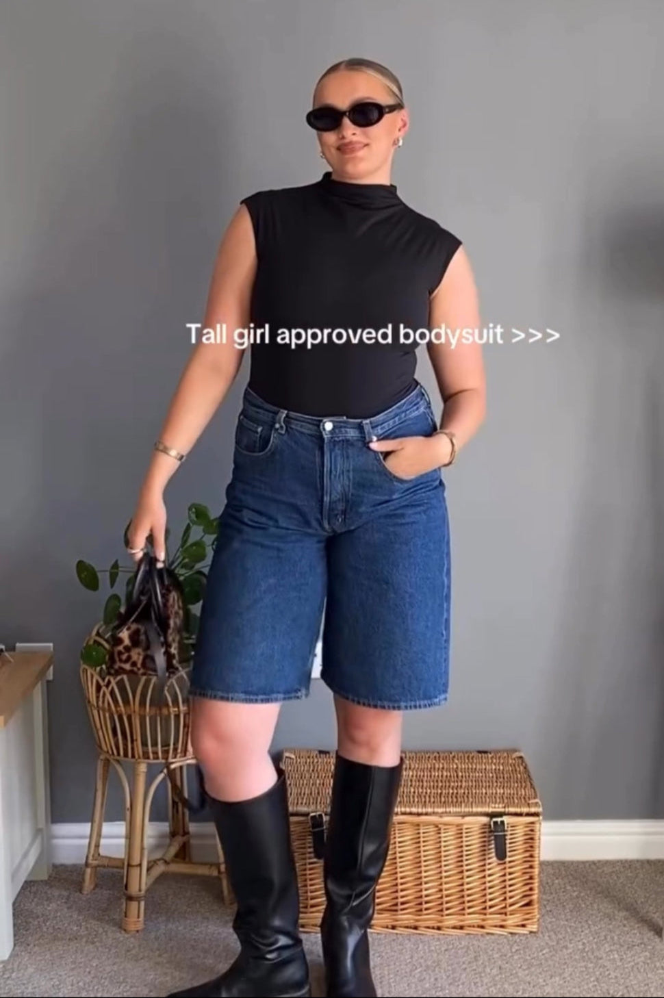 Person wearing a black sleeveless top and blue denim shorts standing against a gray wall.